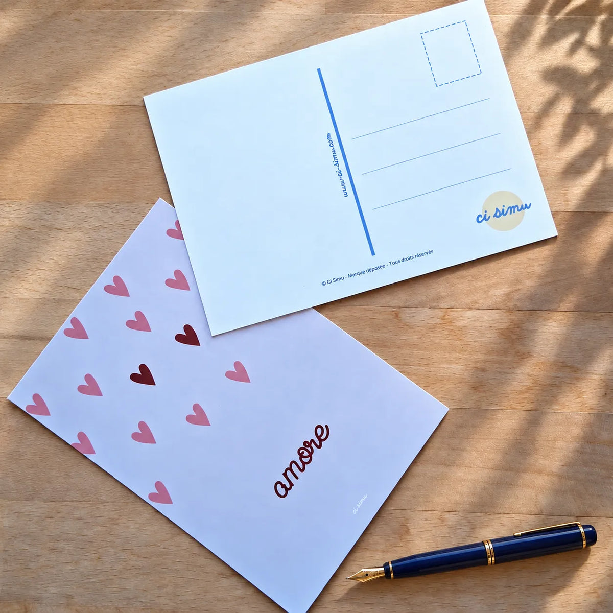 Dos d'une carte postale personnalisée avec motif cœurs « amore » et stylo bille posés sur une table en bois